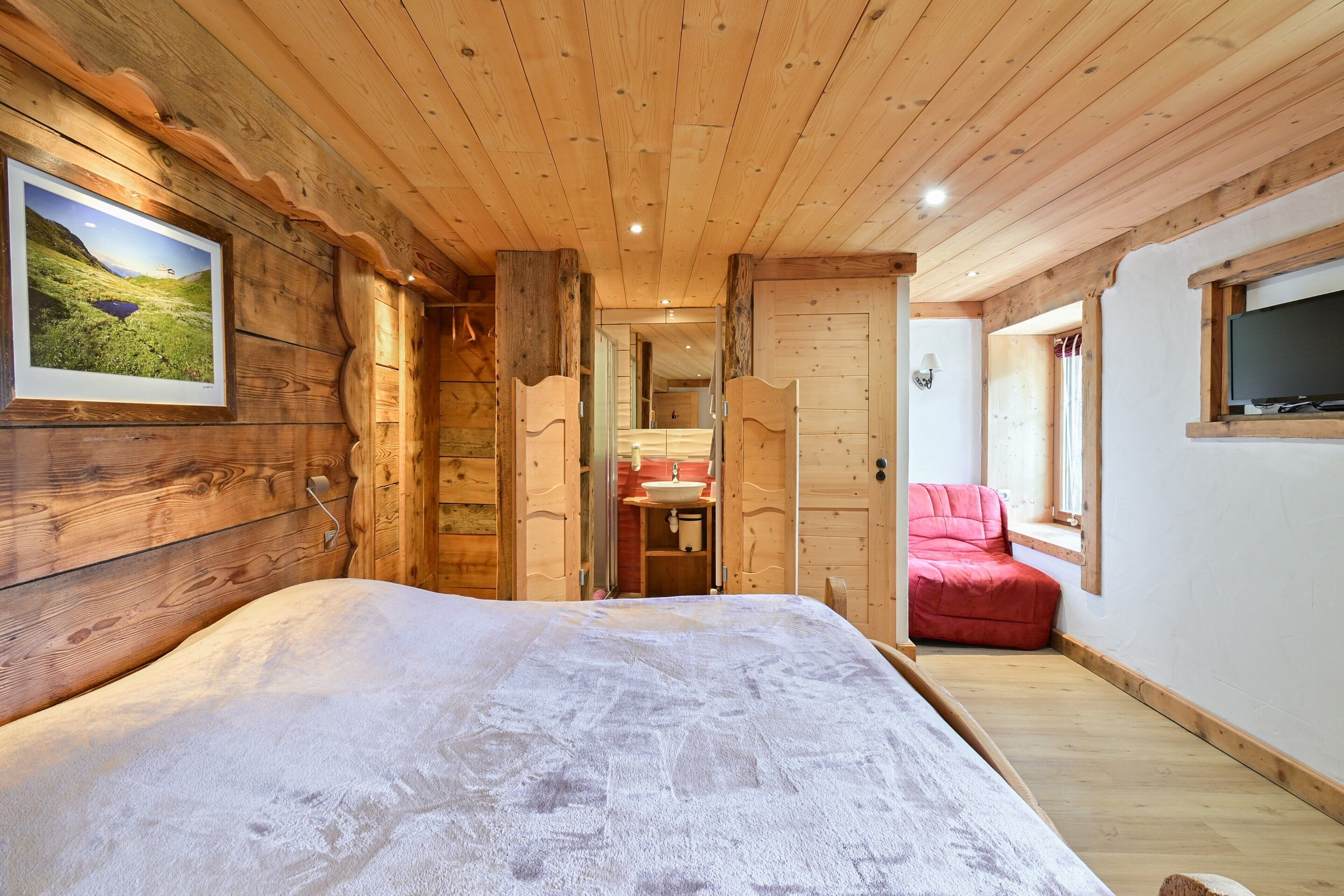A wooden cabin interior with a bed, a red chair, a TV, and a bathroom with a sink.
