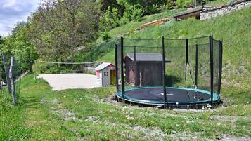 Un trampolín rodeado de una red de seguridad, situado en un césped con un cobertizo pequeño y un muro de piedra al fondo.