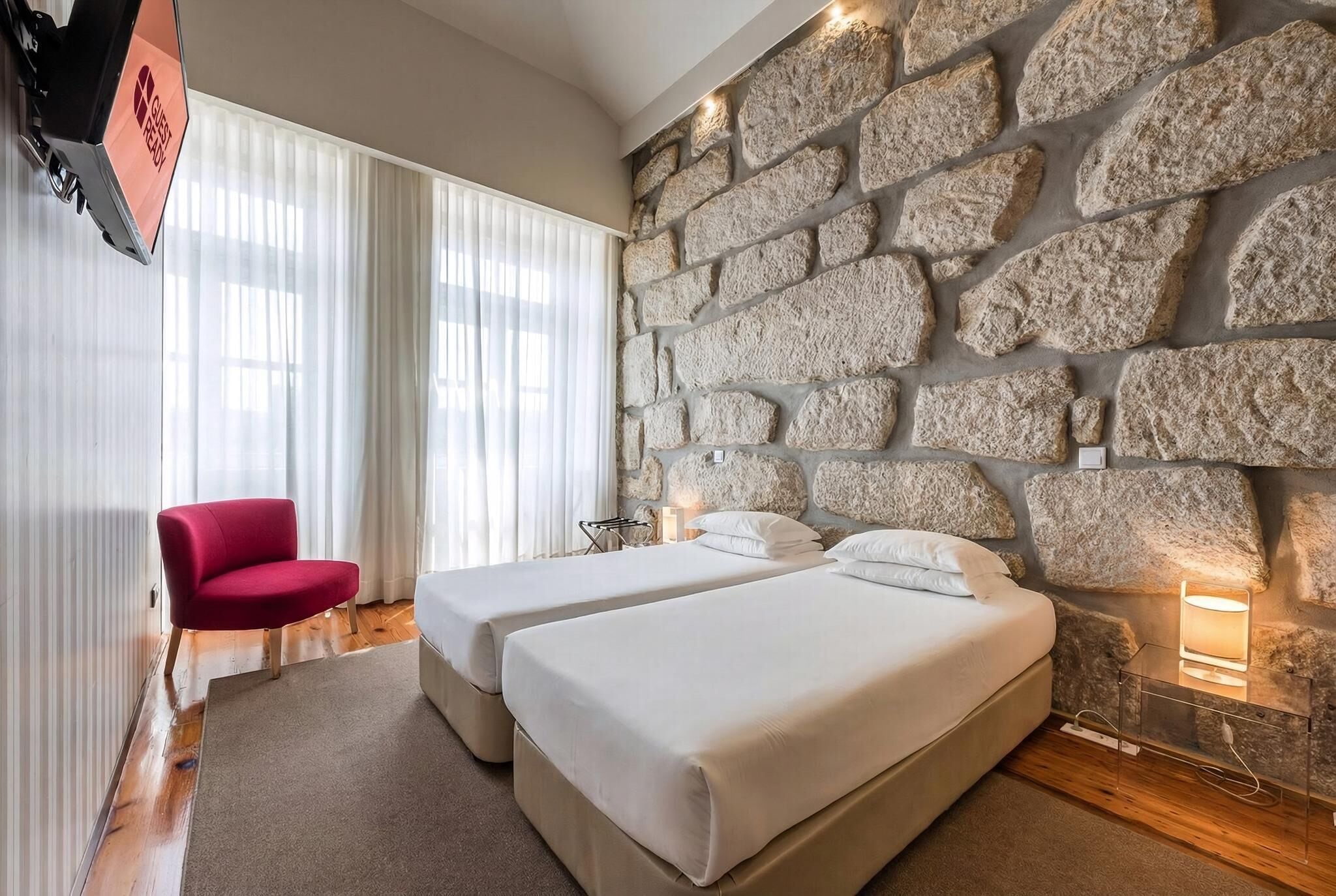 Une chambre d’hôtel avec un mur en pierre, deux lits, un fauteuil rouge et un téléviseur fixé au mur.