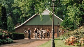 A group of people riding bicycles on a paved path in front of a rustic barn-like structure surrounded by lush greenery. - Cozy vacation homes and apartments - here you will find peace and relaxation (Drantum)