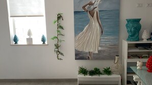 Een kamer met een groot raam, een schilderij van een vrouw aan zee, een bank met planten en een witte plank met decoratieve objecten.