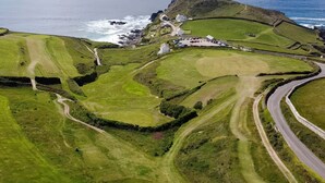 An aerial view of a coastal area with a golf course, a lighthouse, and a small settlement with buildings and a parking area. - Sea Holly (Penzance)