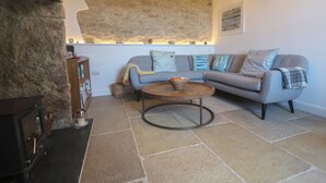 A cozy living room with a stone wall, a grey sofa with patterned cushions, a round wooden coffee table, and a fireplace with a stone mantle. - Sea Holly (Penzance)