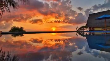 A sunset over a calm body of water with a reflection of the sky, palm trees, and a building with a blue umbrella.