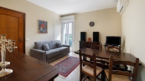 A living room with a sofa, dining table, chairs, a TV, speakers, and a clock on the wall. - Holiday Home 'Casa Bellini' with Private Garden, Wi-Fi and Air Conditioning (Solarolo)