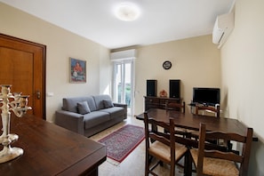 A living room with a sofa, dining table, chairs, a TV, speakers, and a clock on the wall.