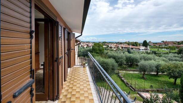 A balcony with a checkered floor, wooden shutters, and a view of a residential area with trees and houses. - Holiday Home 'Casa Bellini' with Private Garden, Wi-Fi and Air Conditioning (Solarolo)