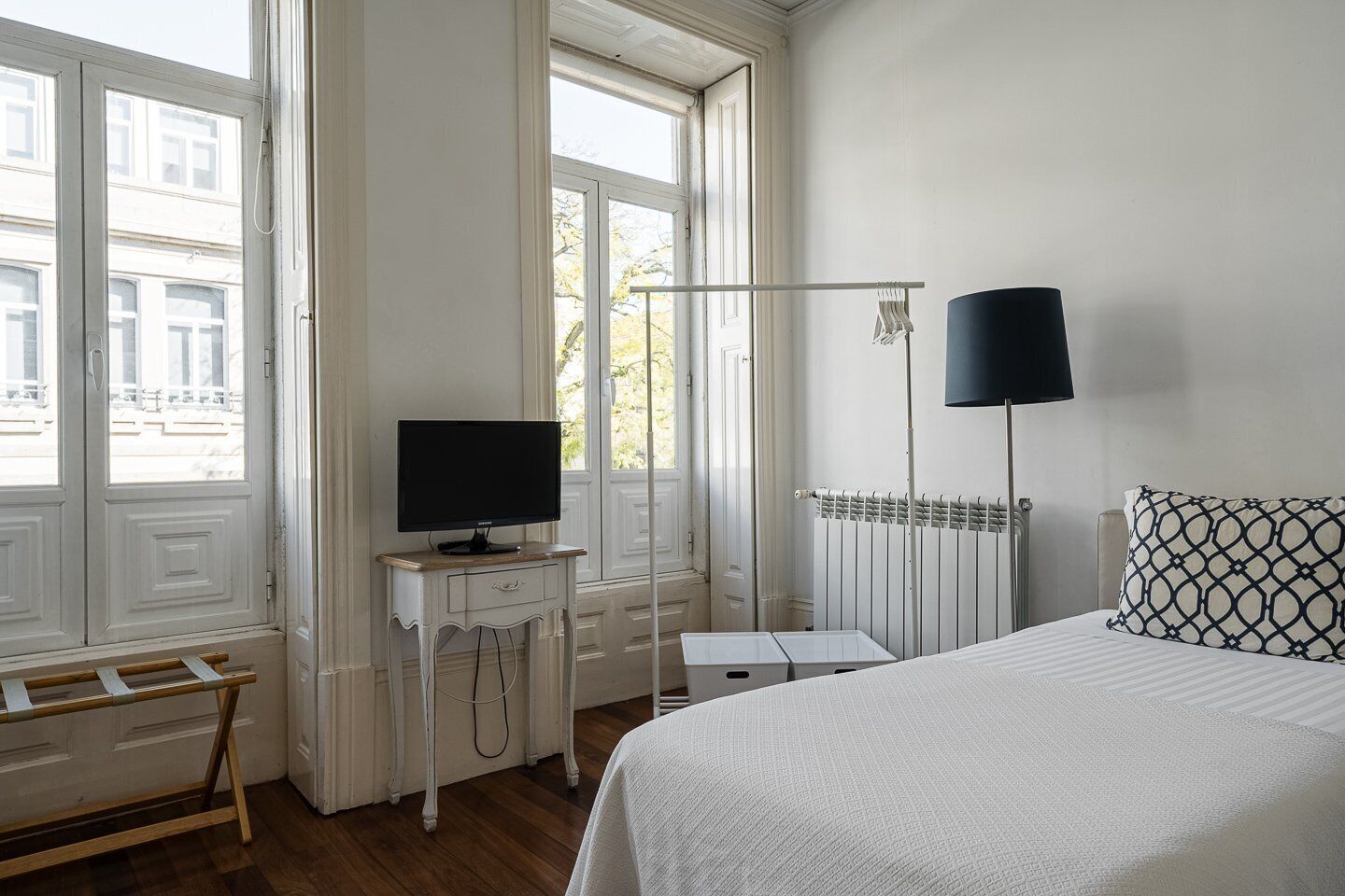 Habitación de hotel con cama, televisión sobre una mesita, radiador y ventana con vistas a edificios.