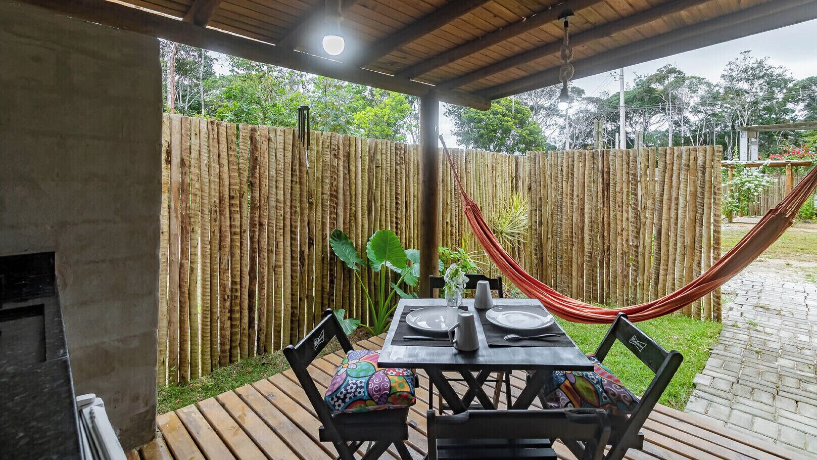 An outdoor patio with a wooden table set for two, surrounded by chairs with colorful cushions. A hammock is draped over the fence, and a potted plant is visible in the background.