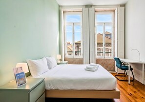 A hotel room with a bed, a desk, a chair, a nightstand with a lamp, and a window with a view of buildings. - Guestready - Porto Home Guesthouse (Porto)