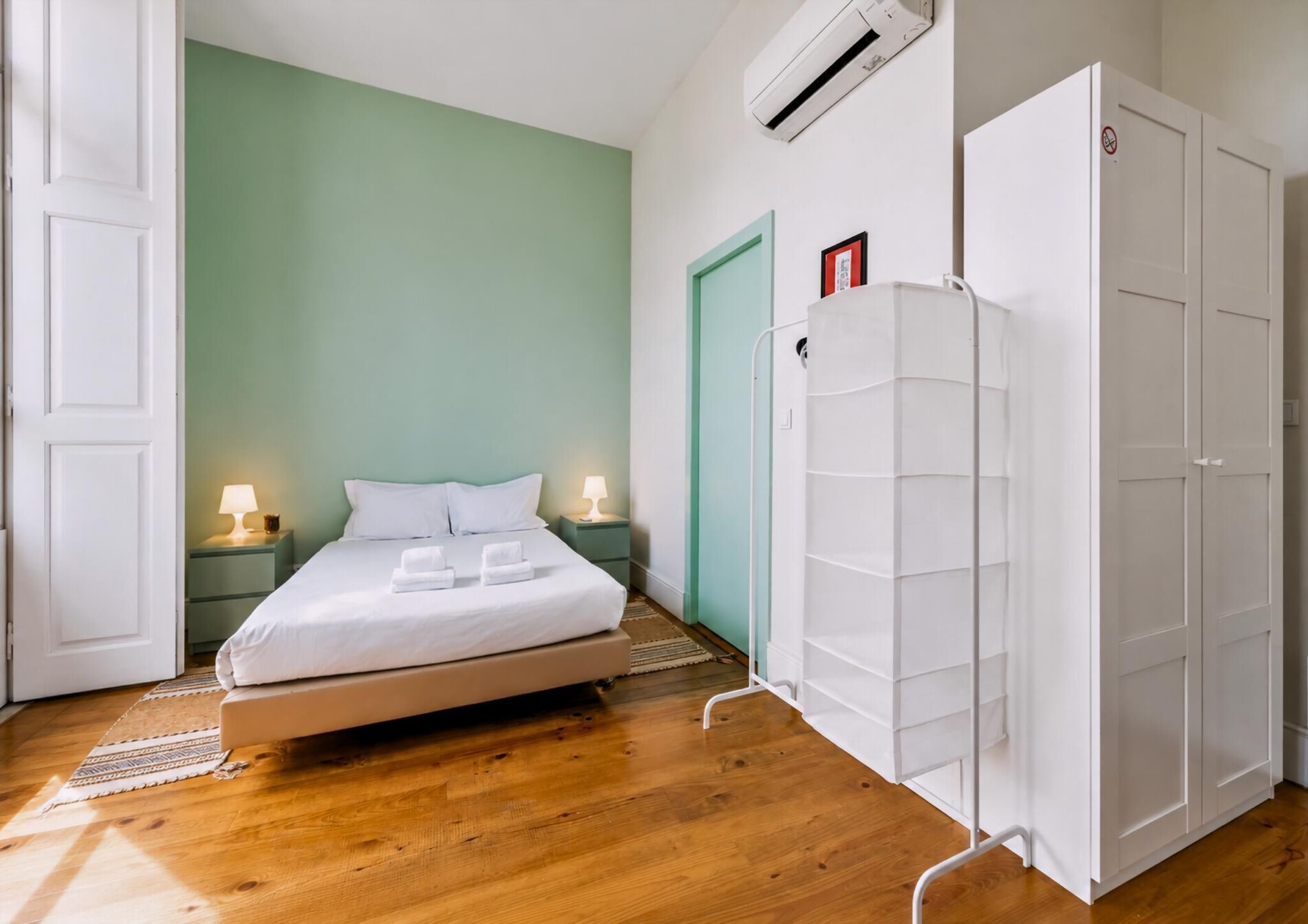 A hotel room with a bed, bedside lamps, a wardrobe, and an air conditioning unit.