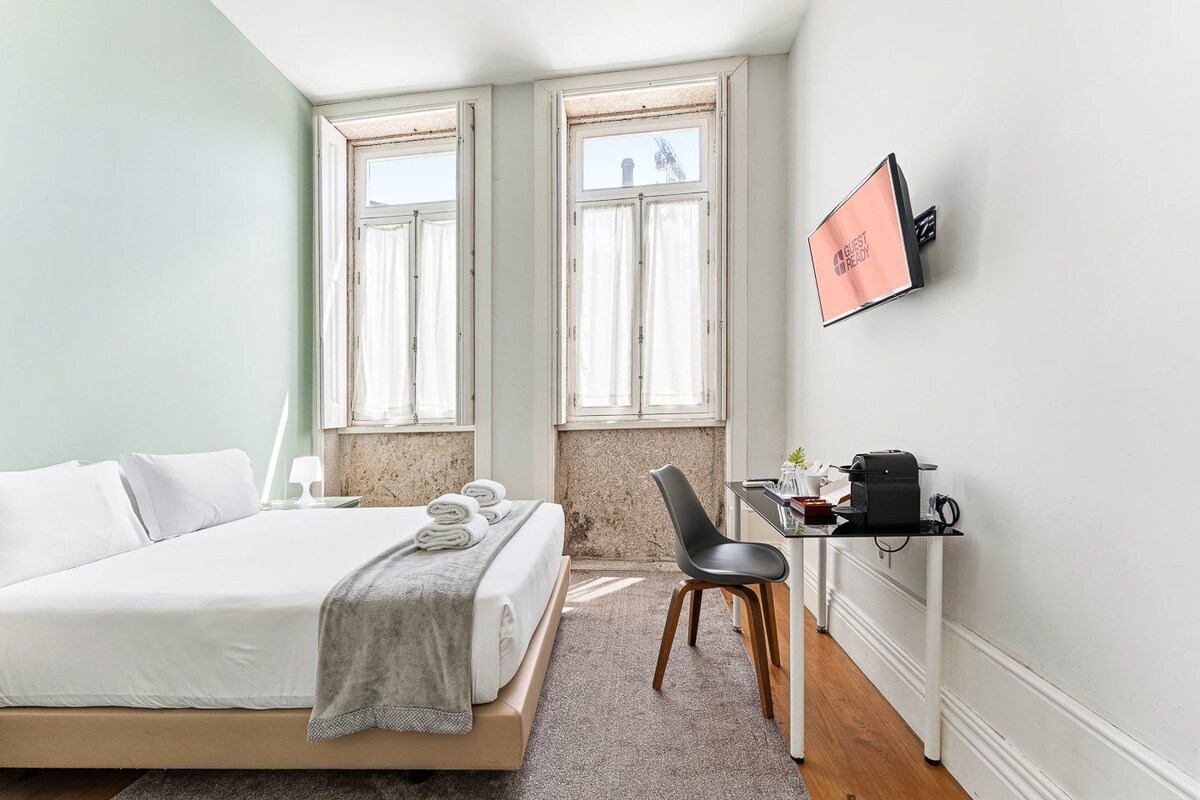 A hotel room with a bed, two windows, a desk with a chair, and a flat-screen TV.