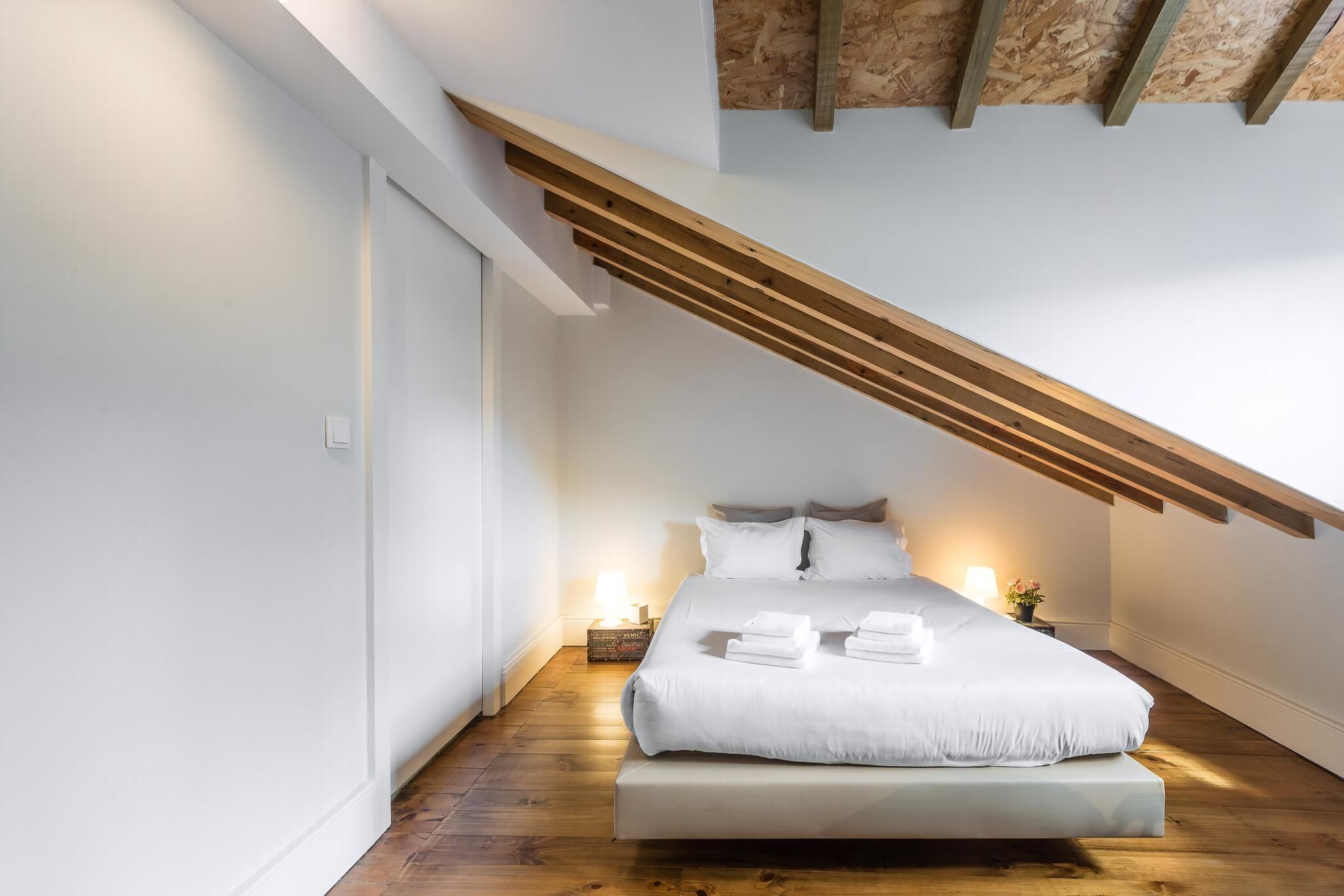 Um quarto moderno com teto inclinado, uma cama grande, luminárias de cabeceira e uma pequena mesa com uma planta.