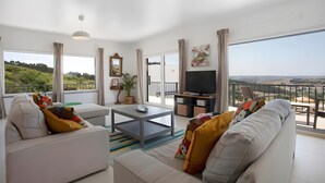 Una sala de estar moderna con sofá, mesa de centro, televisor y ventanas amplias con vistas al paisaje.