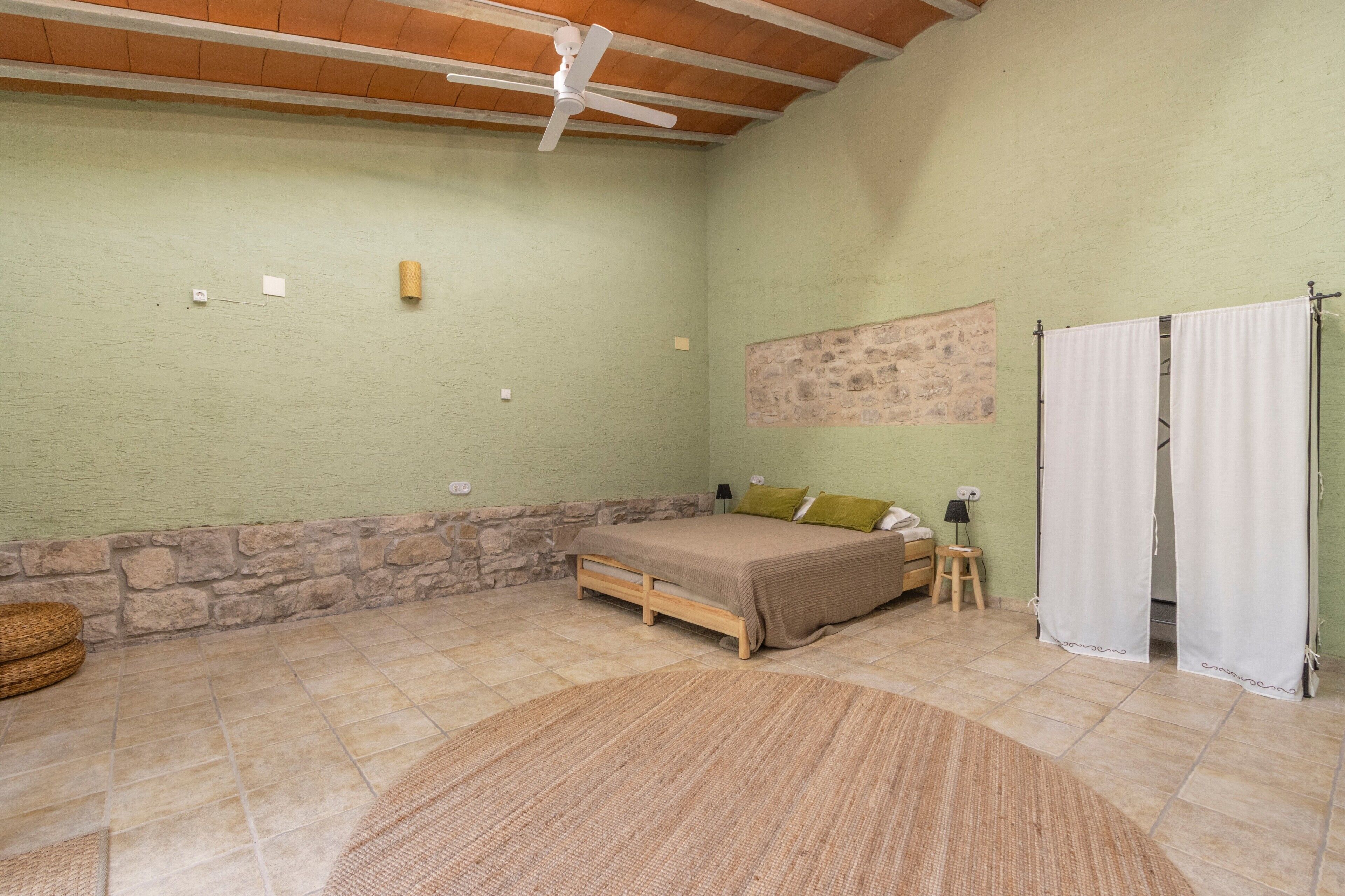 Un dormitori amb llit, ventilador de sostre, paret de pedra, estova i finestra amb cortines.
