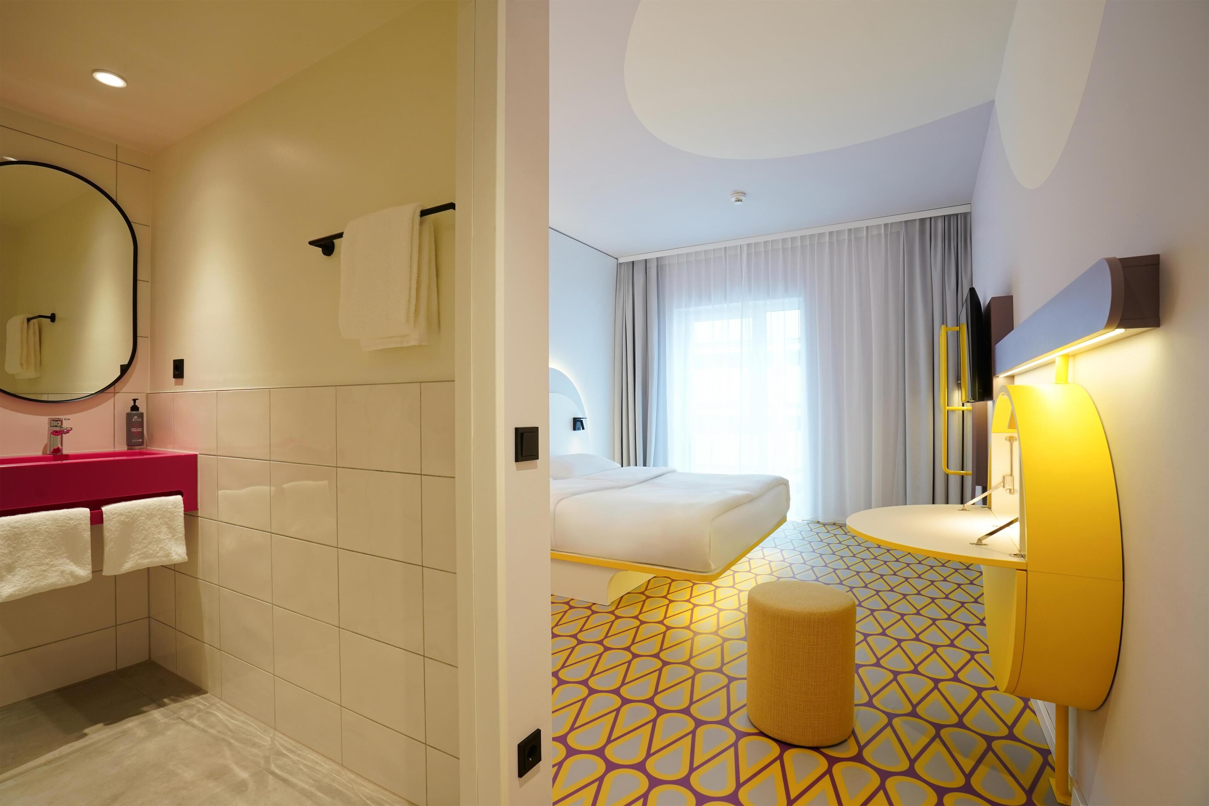 A modern hotel room with a bathroom featuring a pink vanity, a large mirror, and a towel rack. The bedroom has a bed with a white headboard, a bedside table, and a large window with curtains.