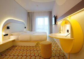 Una habitación de hotel moderna con una cama grande, una mesita de noche, un pequeño taburete, un escritorio curvo y un ventanal con cortinas.