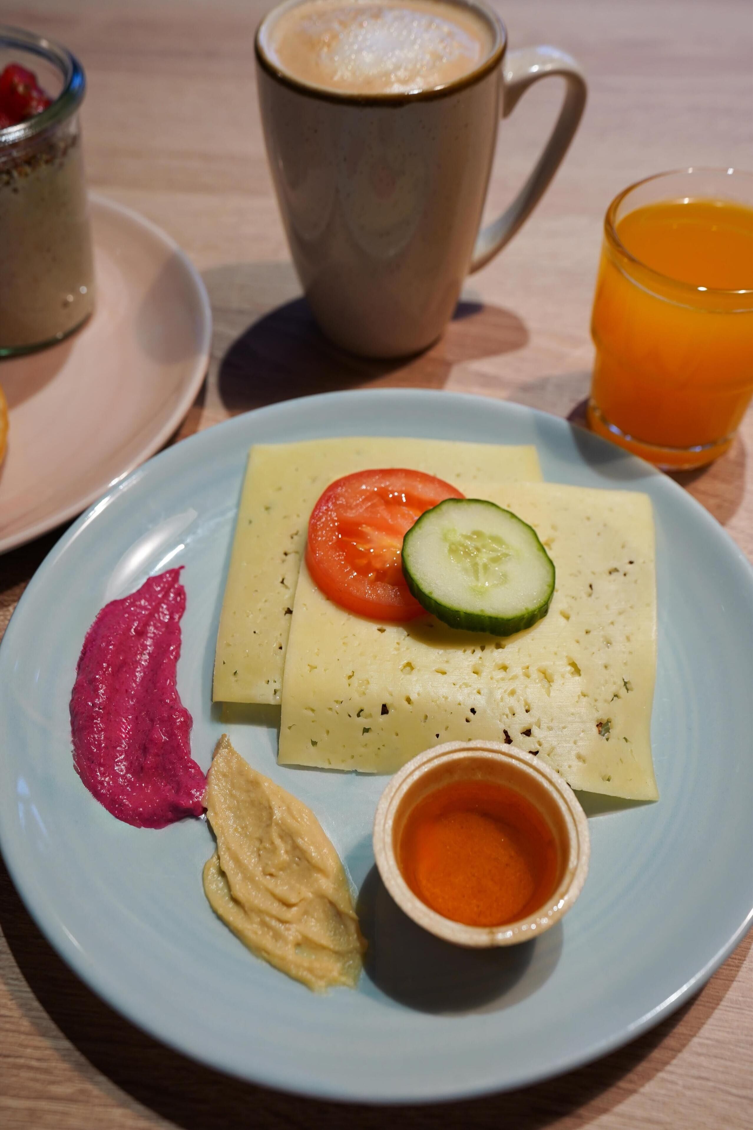Un plato con un sándwich, una rodaja de tomate, un pepino, una cucharada de salsa roja, una cucharada de salsa amarilla y una taza pequeña de salsa naranja.