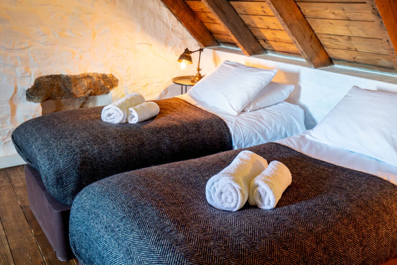 A cozy room with two beds, each with a rolled towel, a lamp, and a rustic stone wall.