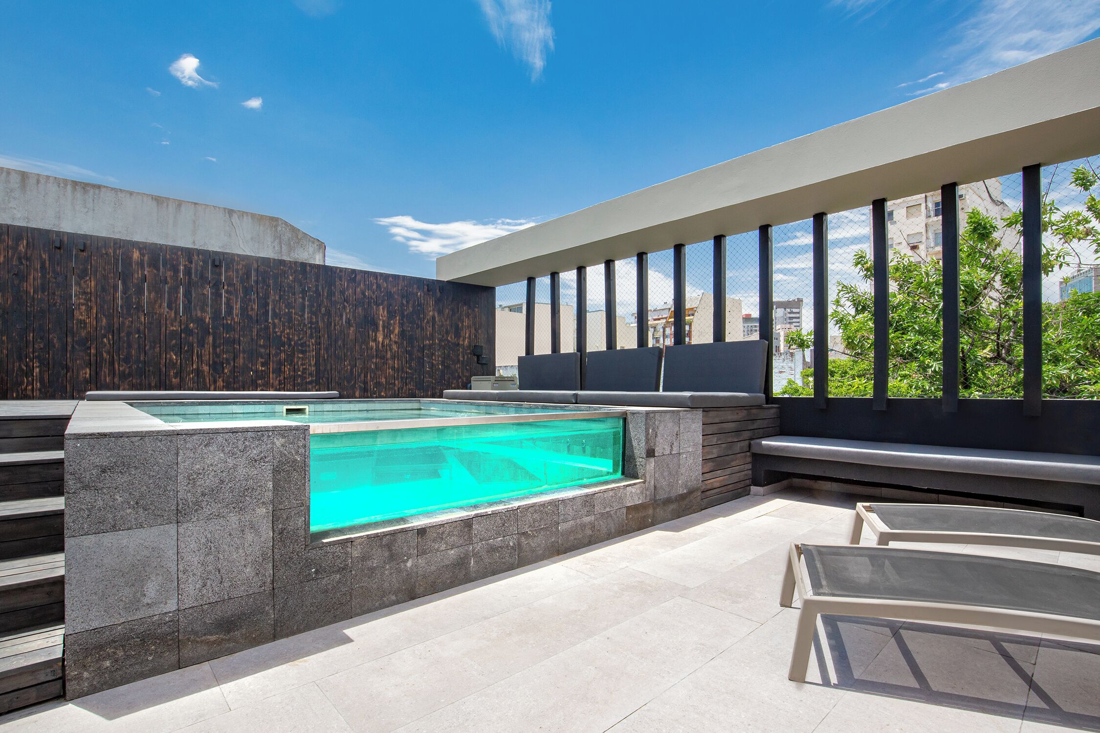 A rooftop pool area with a clear view of the city skyline, a modern design with stone and glass elements, and a seating area with loungers.