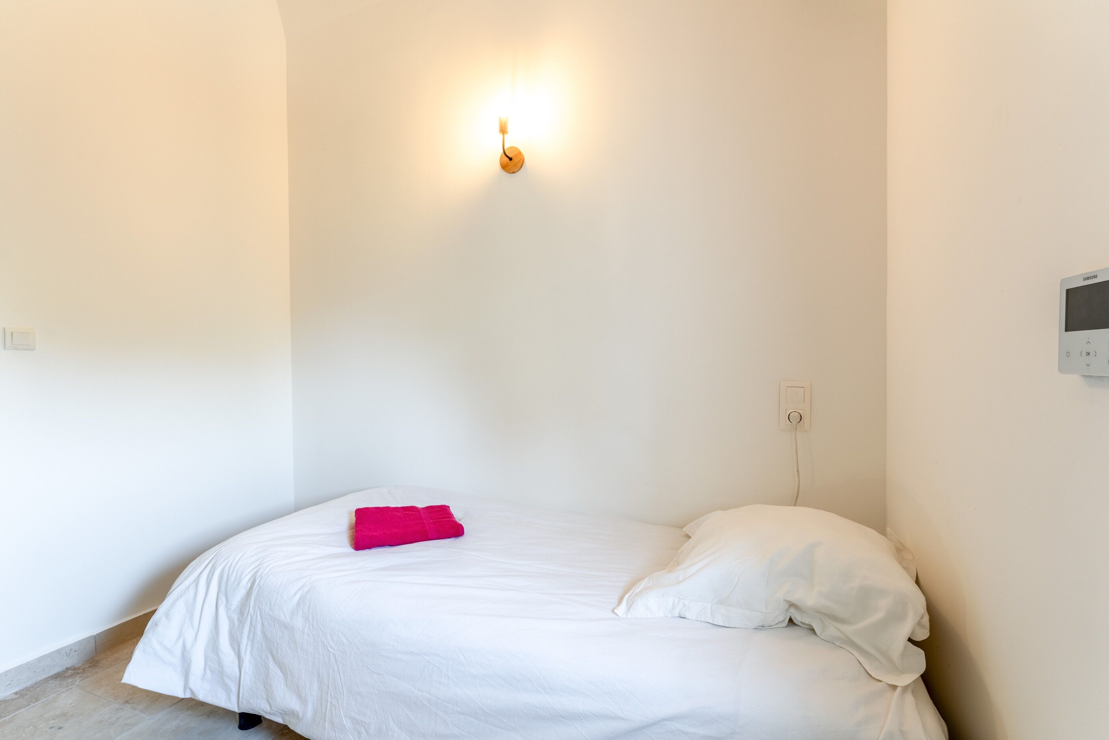 Uma cama de solteiro com roupa de cama branca e um travesseiro rosa, luminária fixada na parede e termostato na parede.