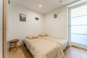 Une pièce avec deux lits, un petit tabouret, une porte et un mur orné d’un tableau et d’un motif représentant un arbre.