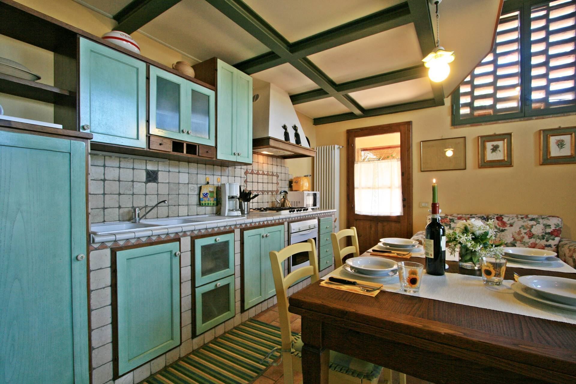 A kitchen with teal cabinets, a tiled backsplash, a stove, and a dining area with a table set for two.