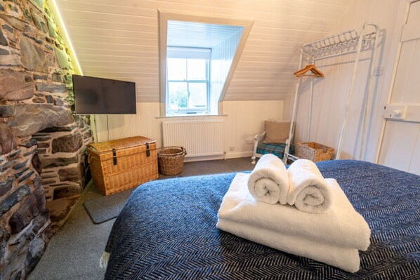 A cozy bedroom with a stone wall, a large window, a bed with rolled towels, a TV, and a hanging rack. - Plockton Sea View Apartment (Highland)