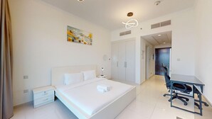 A modern hotel room with a large bed, a desk, and a chair. There is a painting on the wall and a door leading to another room.