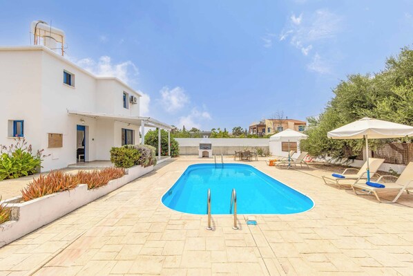 A swimming pool with two ladders, surrounded by lounge chairs and umbrellas, with a white building and greenery in the background.