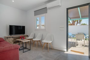 A modern living room with a flat-screen TV, a red sofa, a small table with flowers, and three white chairs.