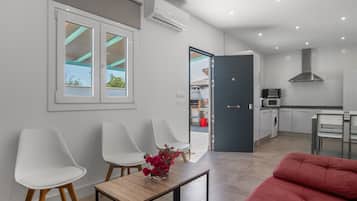 A modern living room with a sofa, two white chairs, a small table with a vase, and a view to a kitchen area.