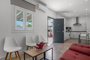 A modern living room with a sofa, two white chairs, a small table with a vase, and a view to a kitchen area.