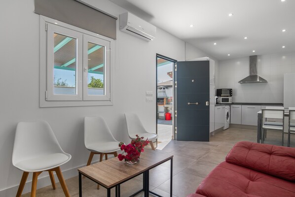 Un salon moderne avec un canapé, deux fauteuils blancs, une petite table sur laquelle est posé un vase, et une vue sur l’espace cuisine.