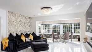 Una sala de estar moderna con un sofá modular negro, una araña y un comedor con vistas amplias a través de grandes ventanales.