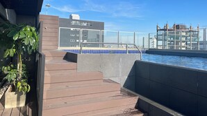 Ett poolområde på taket med en modern byggnad i bakgrunden, en trappa som leder ner till poolen och en krukväxt till vänster.