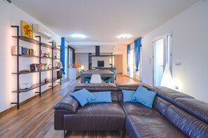 A modern living room with a leather sofa, a bookshelf, a dining area, and a kitchen in the background.