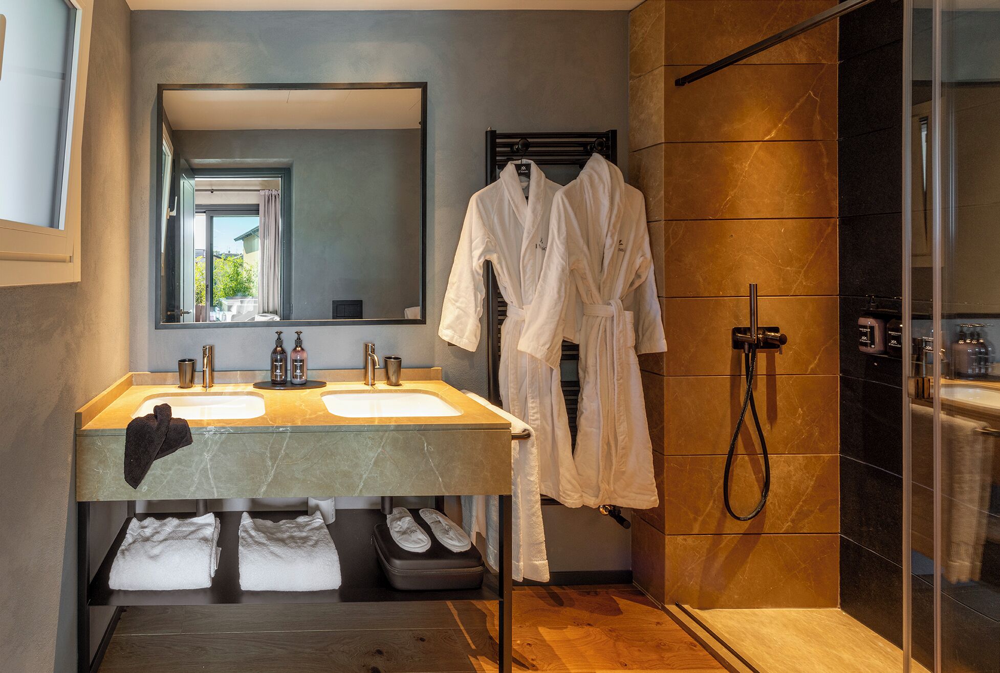 Un baño moderno con lavamanos dobles, un espejo grande, cabina de ducha con mampara de vidrio y batas blancas colgadas en un perchero.