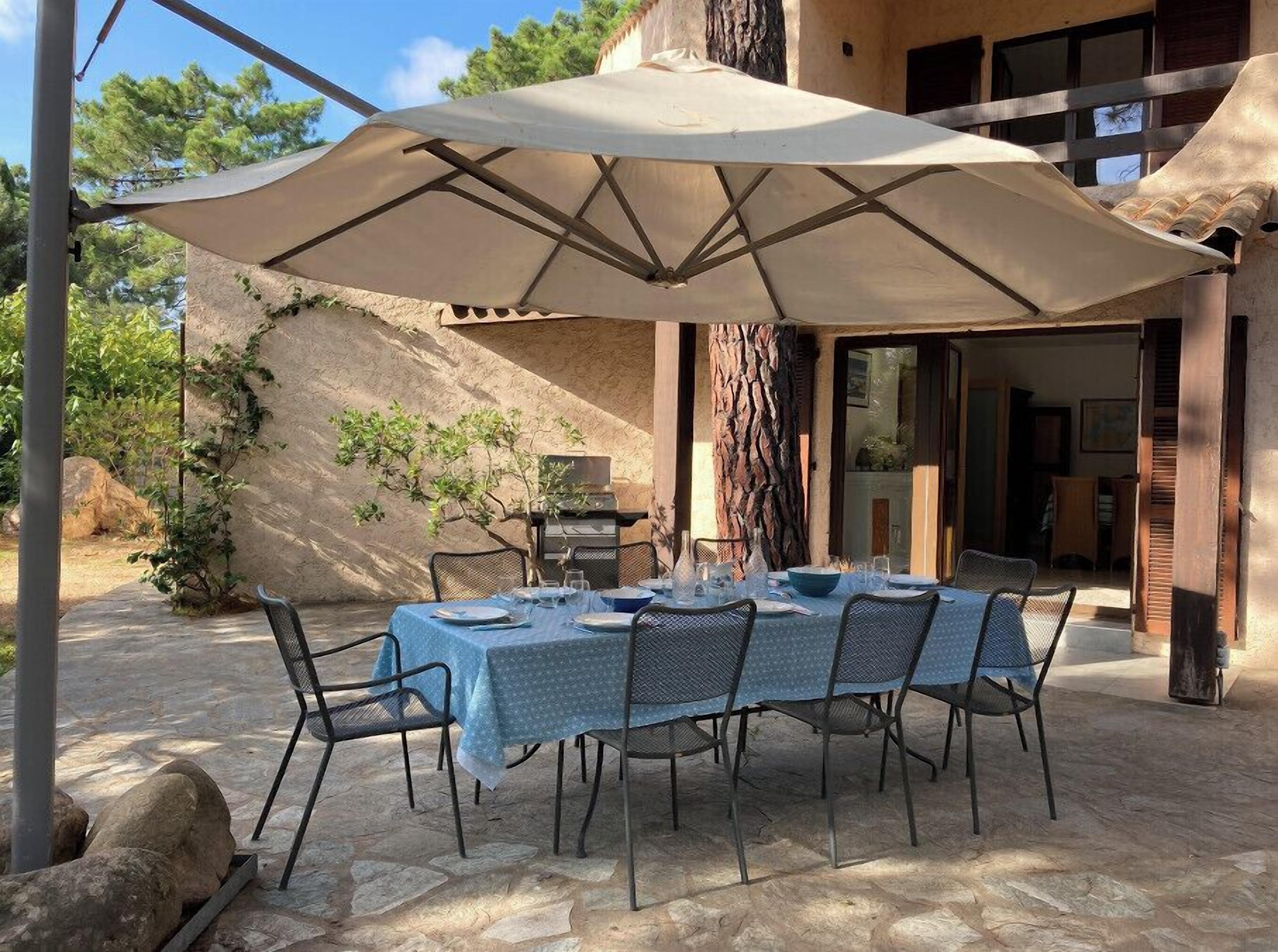 Un área de comedor al aire libre con una mesa dispuesta para seis personas, rodeada de sillas, bajo un paraguas grande.