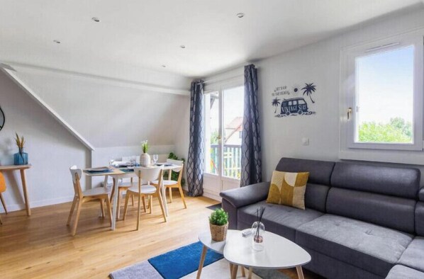 Un saló modern amb un sofà gris, una taula de dinar amb caires, una taula petita blanca i una escala que mena a un pis superior.