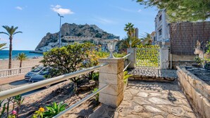A coastal road with a stone pathway, palm trees, and a view of the sea and a mountain.