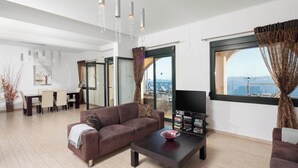 Una sala de estar moderna con un sofá marrón, una mesita de centro y un televisor. Hay un comedor con una mesa y sillas, y vistas al mar a través de amplios ventanales.
