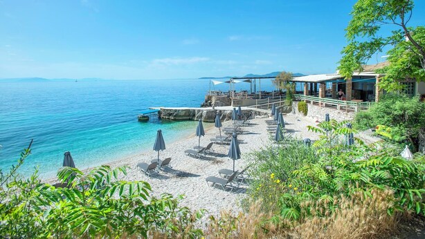 Una playa costera con un restaurante con vistas al mar, tumbonas y un litoral rocoso.
