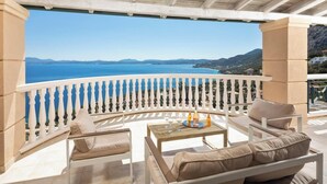 Un balcón con vista al mar, una mesa con vasos y botellas, y asientos acolchados color beige.