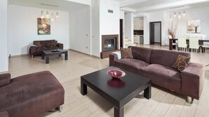 Una sala de estar moderna con un sofá marrón, una mesa de centro negra y una chimenea. Se observa un comedor con una mesa y sillas blancas al fondo.