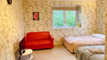 A room with floral wallpaper, a red sofa, a bed with a green bedspread, and a window with a view of greenery.