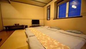 A hotel room with a bed, a TV, a small table, and a window with blue curtains. - Miles Town (Ito)