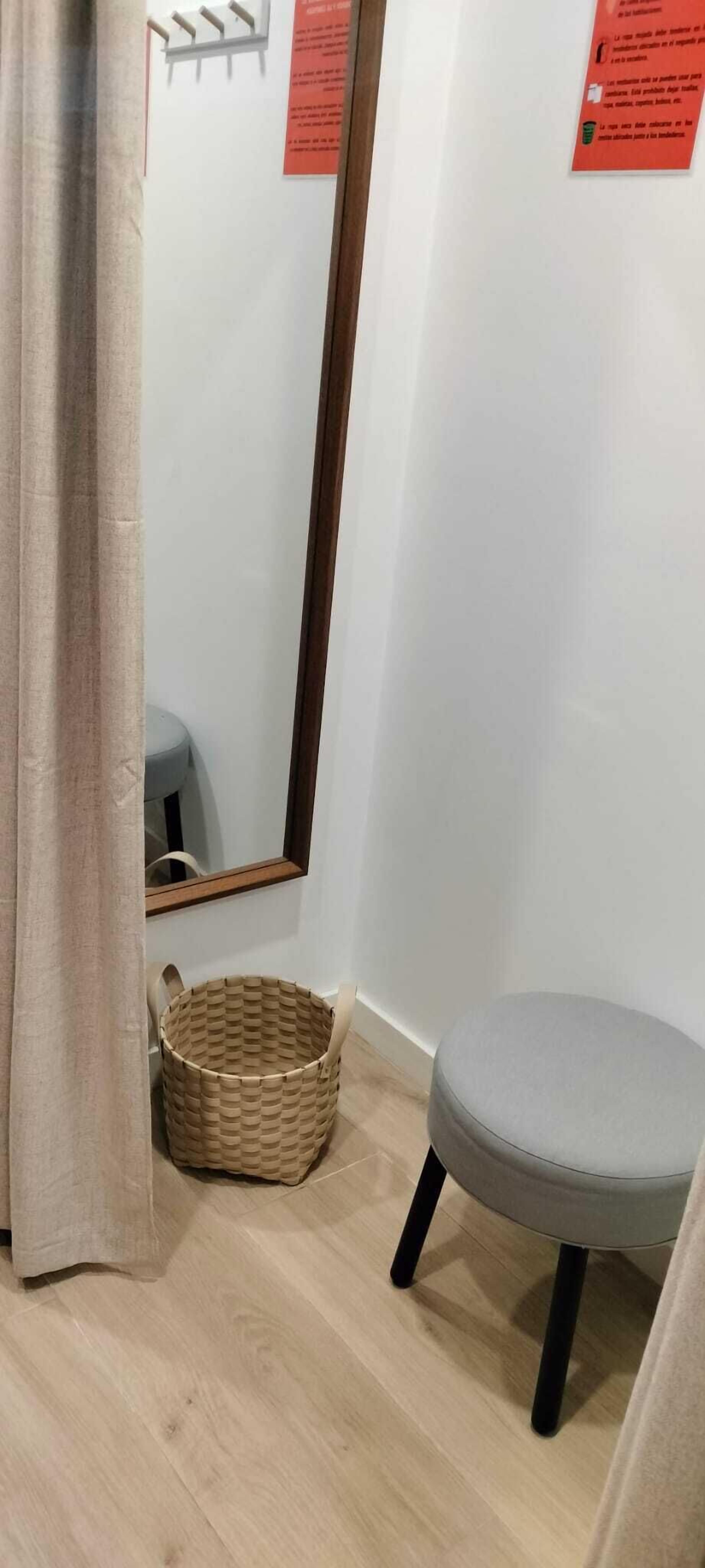Un rincón de una habitación con un taburete acolchado gris, una cesta tejida y un espejo en la pared.