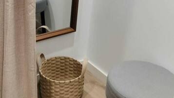 Un rincón de una habitación con un taburete acolchado gris, una cesta tejida y un espejo en la pared.