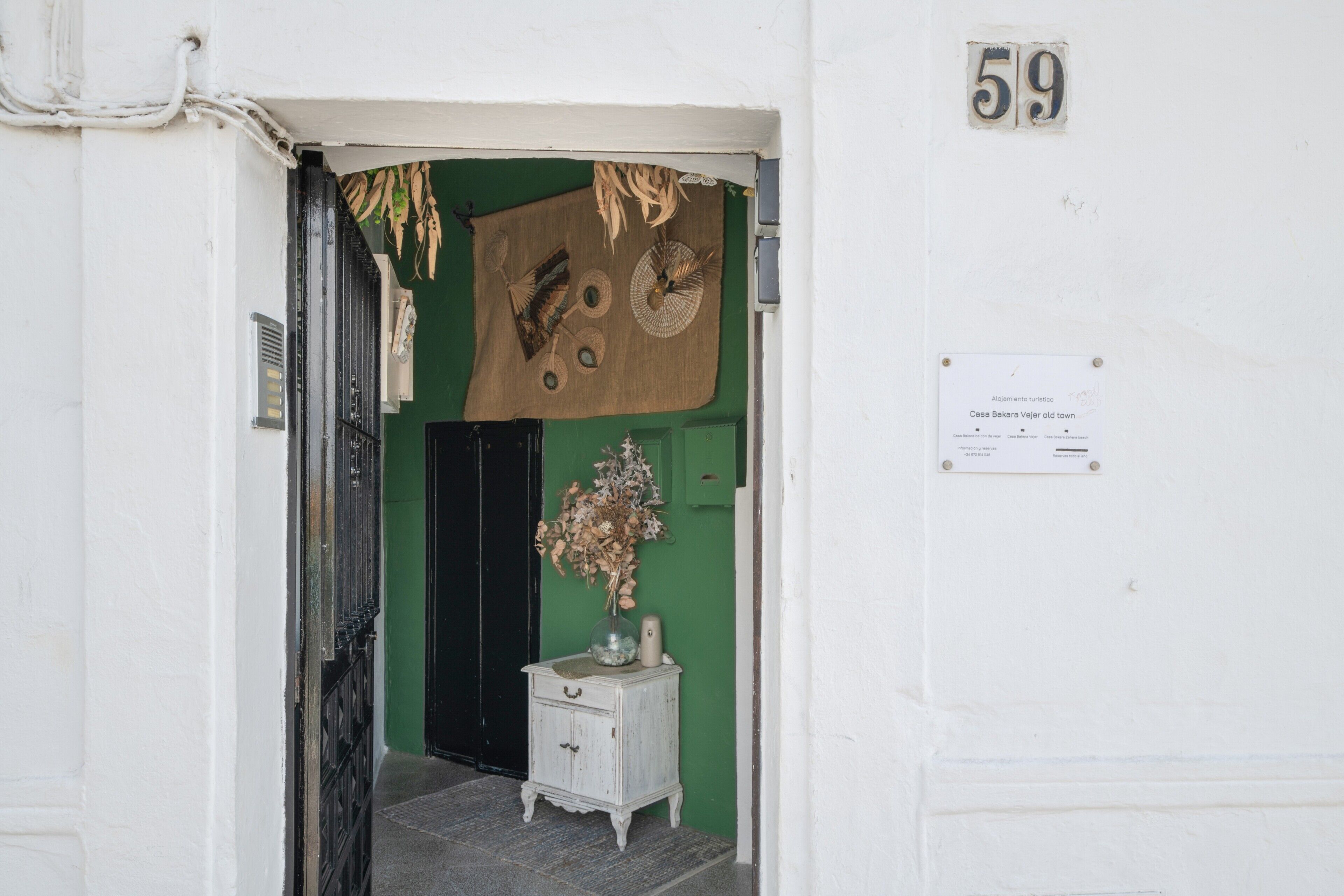 Une entrée d’immeuble blanche avec une porte verte, une plaque portant le numéro « 59 », et une petite table blanche sur laquelle est posé un vase.
